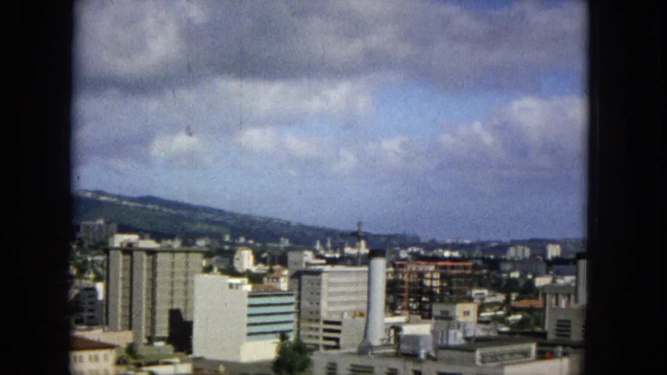 1965 Honolulu overlook showing taller city buildings, a green hillside or ridge, cloudy sky, and foreground rooftops within the dark-edged Super 8 frame.