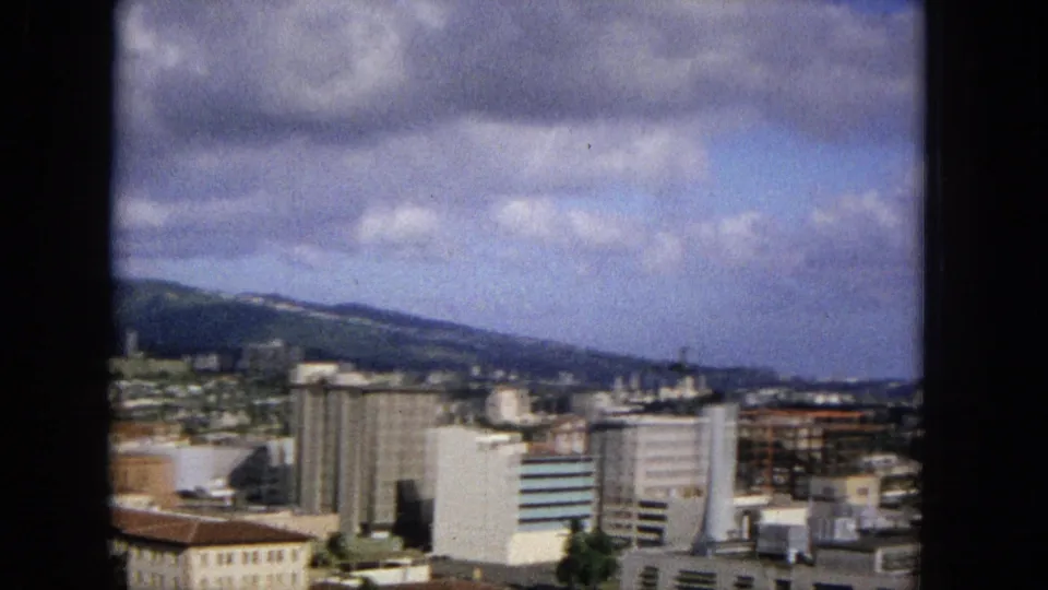 1965 8mm view of a United States city skyline with mid-rise buildings, mountains in the distance, and a cloudy blue sky, framed by dark film borders.