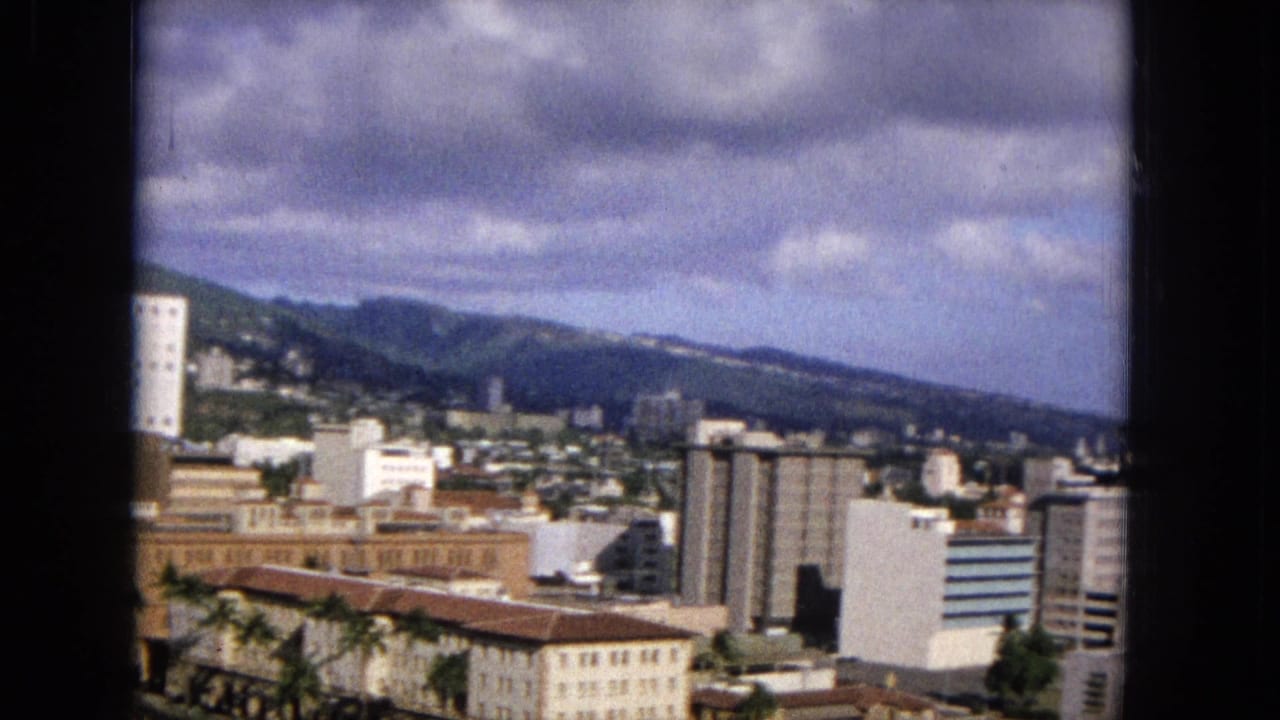 Elevated 1965 8mm city view showing high-rise buildings, a dense urban district, palm trees, mountains, and a cloud-filled sky in the United States.