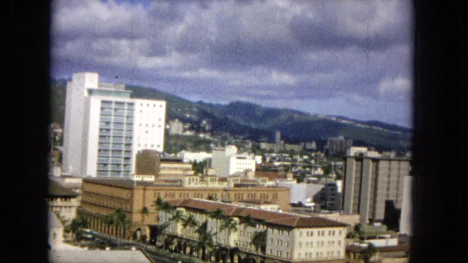1965 8mm frame of a white high-rise, surrounding mid-century city buildings, palm trees, and distant mountains under heavy clouds.