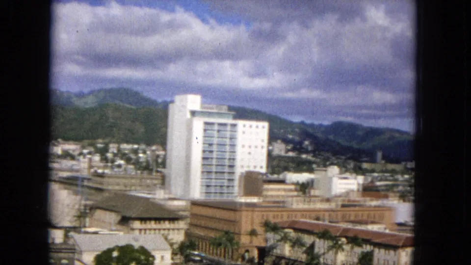 Elevated 1965 United States cityscape with a prominent white tower, palm-lined low buildings, distant green mountains, and a textured cloudy sky.