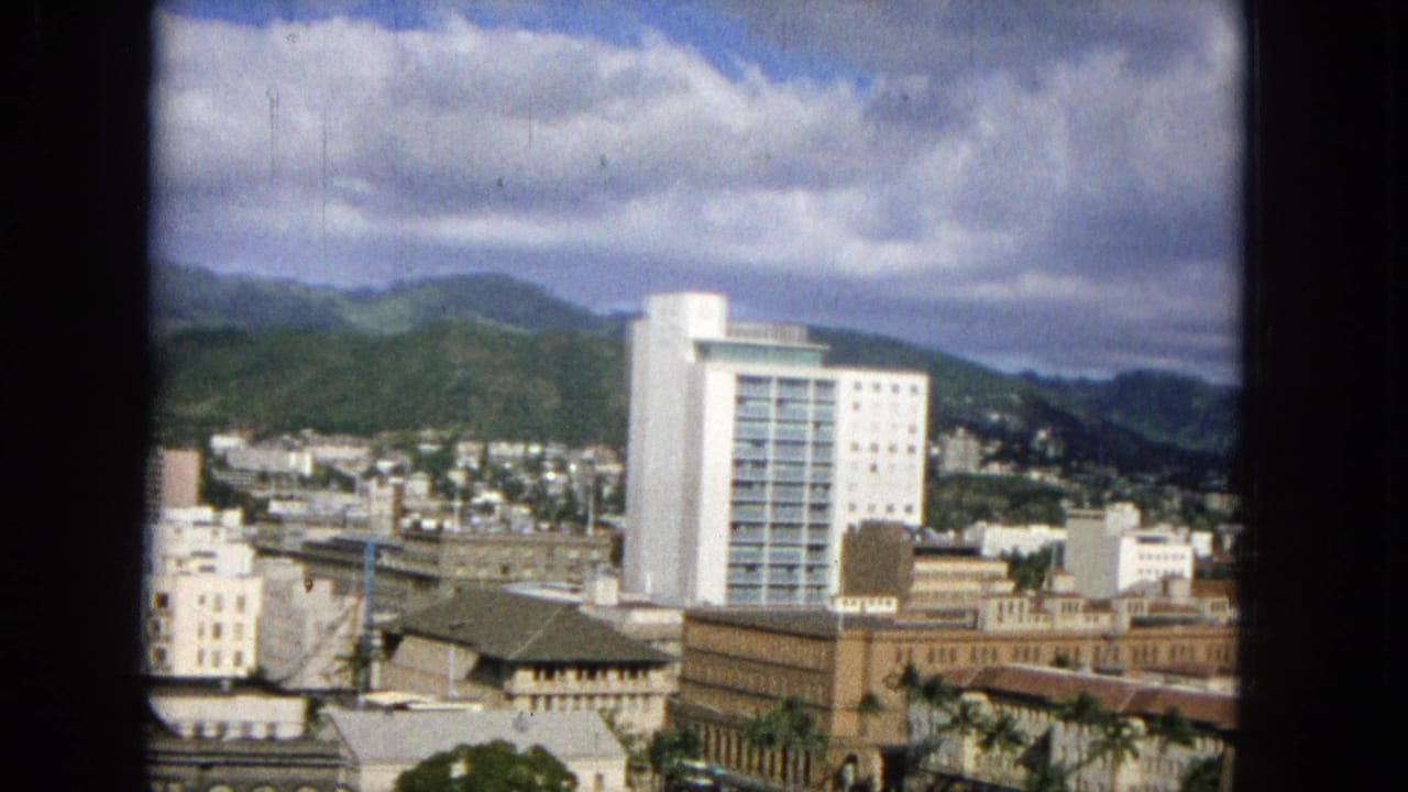 1965 Honolulu city view from an elevated position, showing a prominent white high-rise, surrounding downtown buildings, hillside homes, and cloudy mountains in the background.