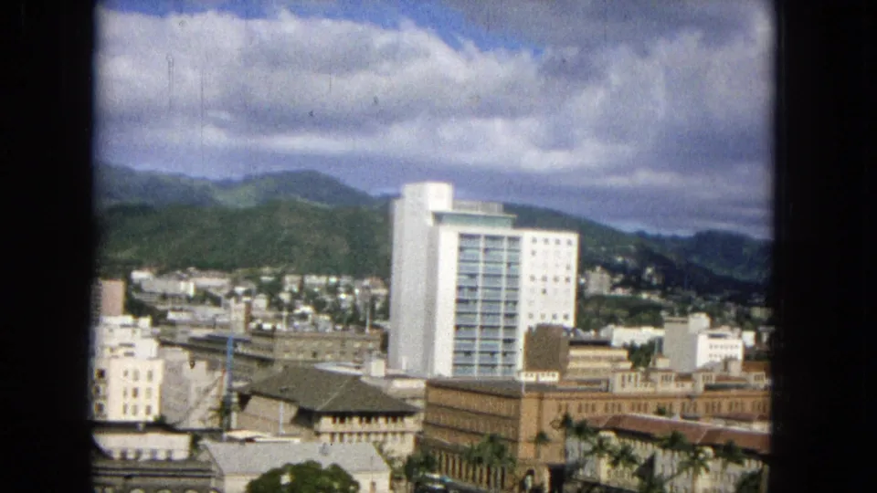 1965 Honolulu city view from an elevated position, showing a prominent white high-rise, surrounding downtown buildings, hillside homes, and cloudy mountains in the background.