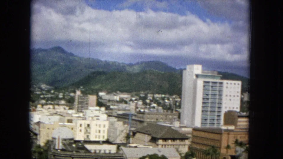1965 Honolulu panorama with a white tower at right, low commercial buildings in the foreground, hillside residences, and green mountain ridges under broken cloud cover.