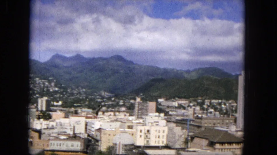 1965 Honolulu cityscape looking toward green mountains, with rooftops, mid-rise buildings, hillside homes, and a partly cloudy sky visible in soft Super 8 color.
