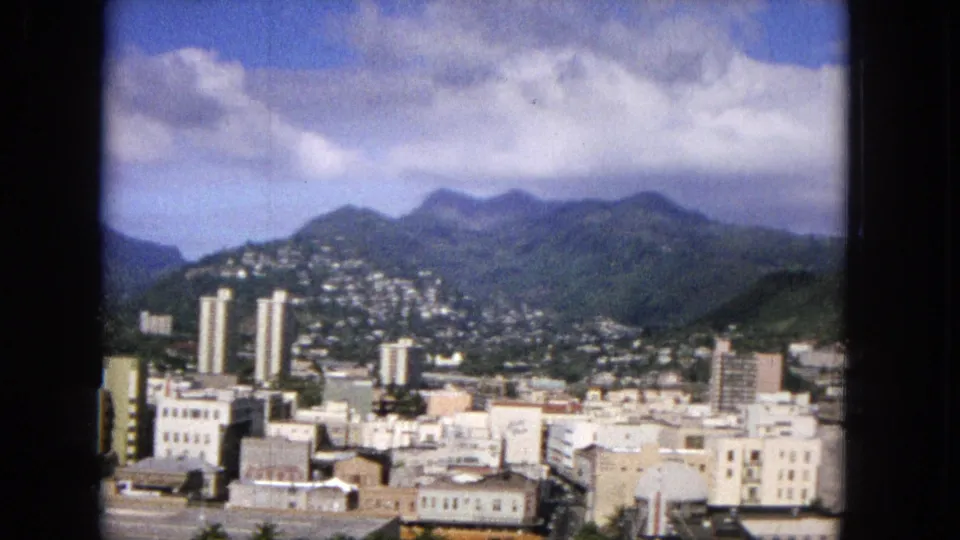 1965 Honolulu view with clustered hillside homes, several taller buildings, downtown rooftops, green mountain slopes, and a partly cloudy sky captured on Super 8 film.