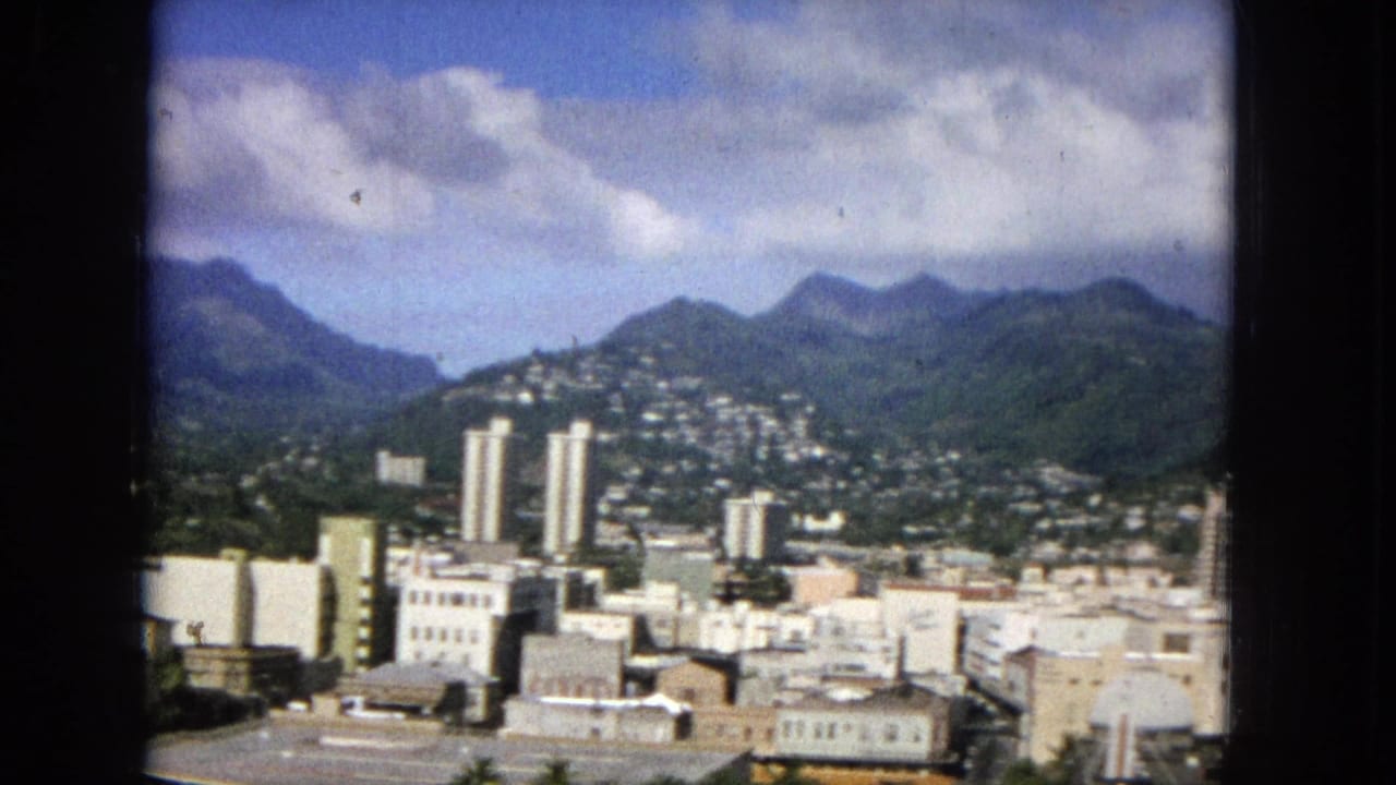 1965 archival 16mm view of a Hawaiian cityscape with high-rise towers, low downtown buildings, hillside residences, green mountains, and blue sky.