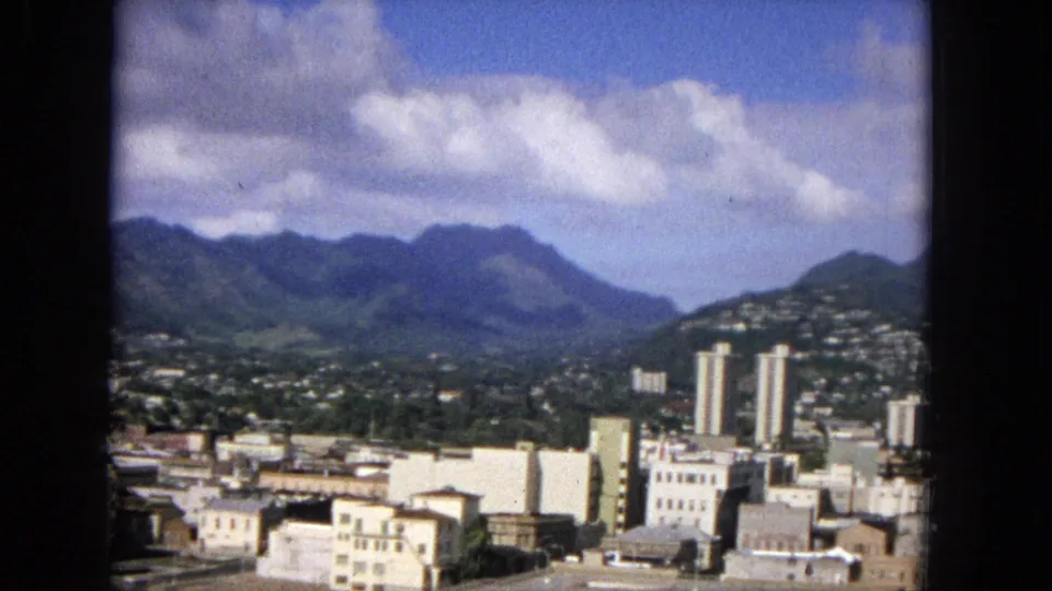 1965 home-movie frame of a Hawaiian urban area with pale buildings, high-rise towers, hillside housing, a green valley, and dark mountains under a clouded sky.
