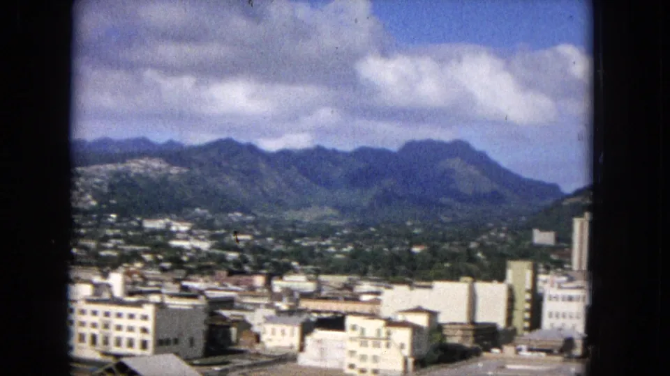 Archival 16mm frame from 1965 showing a wider Hawaiian city-and-mountain panorama with low buildings in the foreground, hillside development, and layered green ridges.