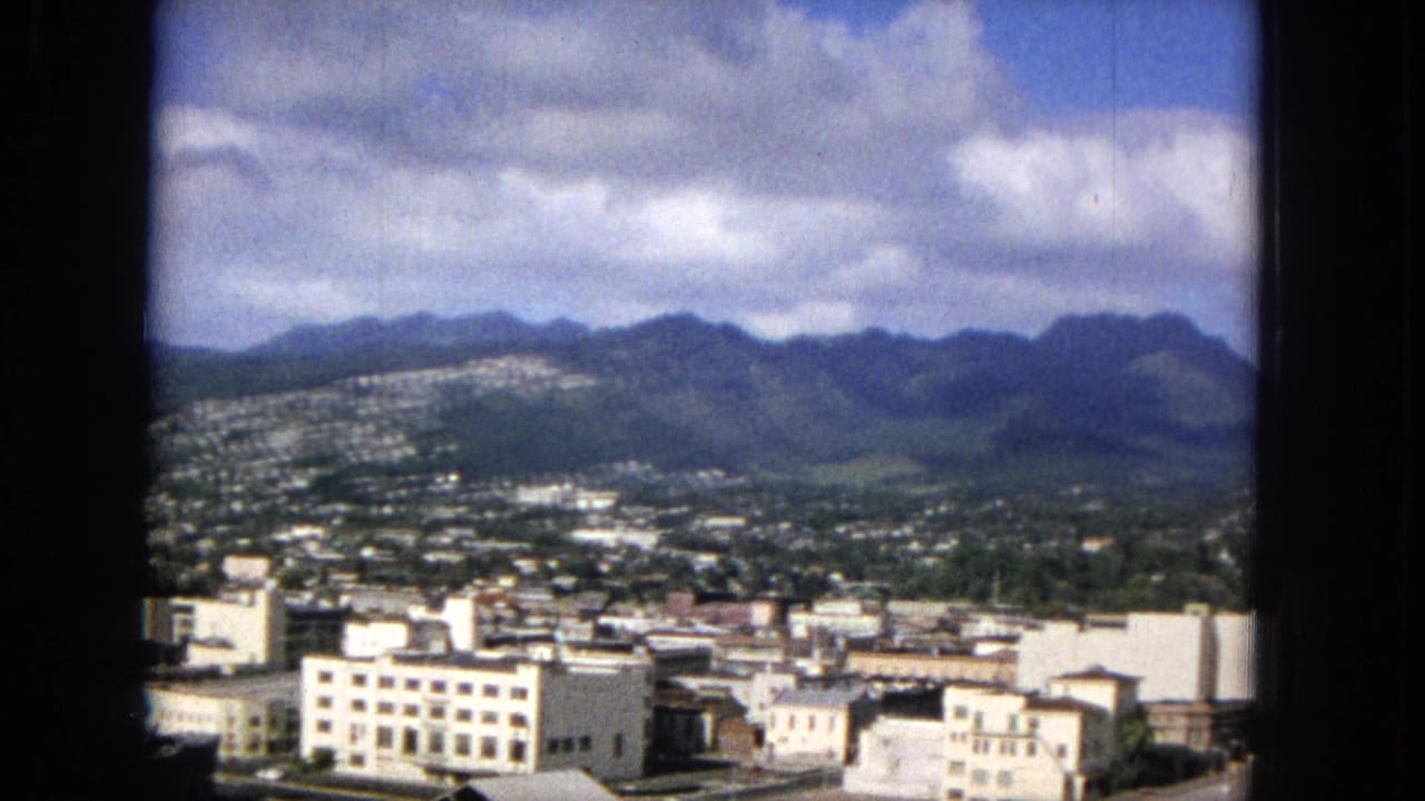 1965 Hawaiian city view, likely Honolulu, showing white downtown buildings below green mountain ridges and scattered clouds, framed by dark film borders.