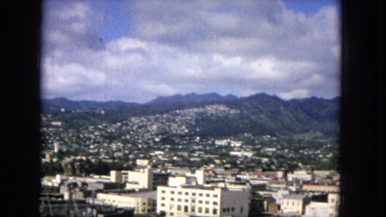 Urban buildings and hillside neighborhoods spread below a mountain ridge and clouded sky.