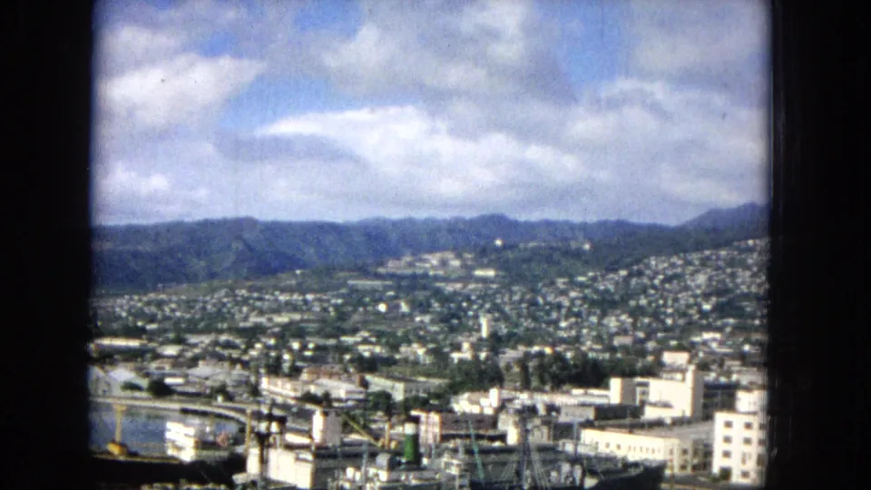 1965 Hawaiian waterfront city view showing harbor activity in the lower frame, urban buildings, hillside housing, and mountain ridges in the distance.