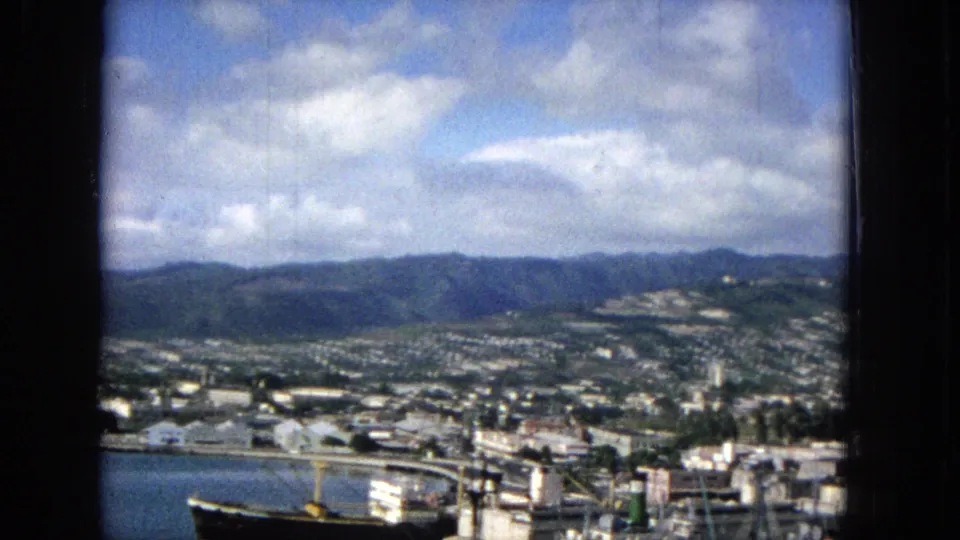 End of the 1965 Hawaii panorama, with harbor water, waterfront cranes or masts, low urban buildings, distant hills, and a bright partly cloudy sky.