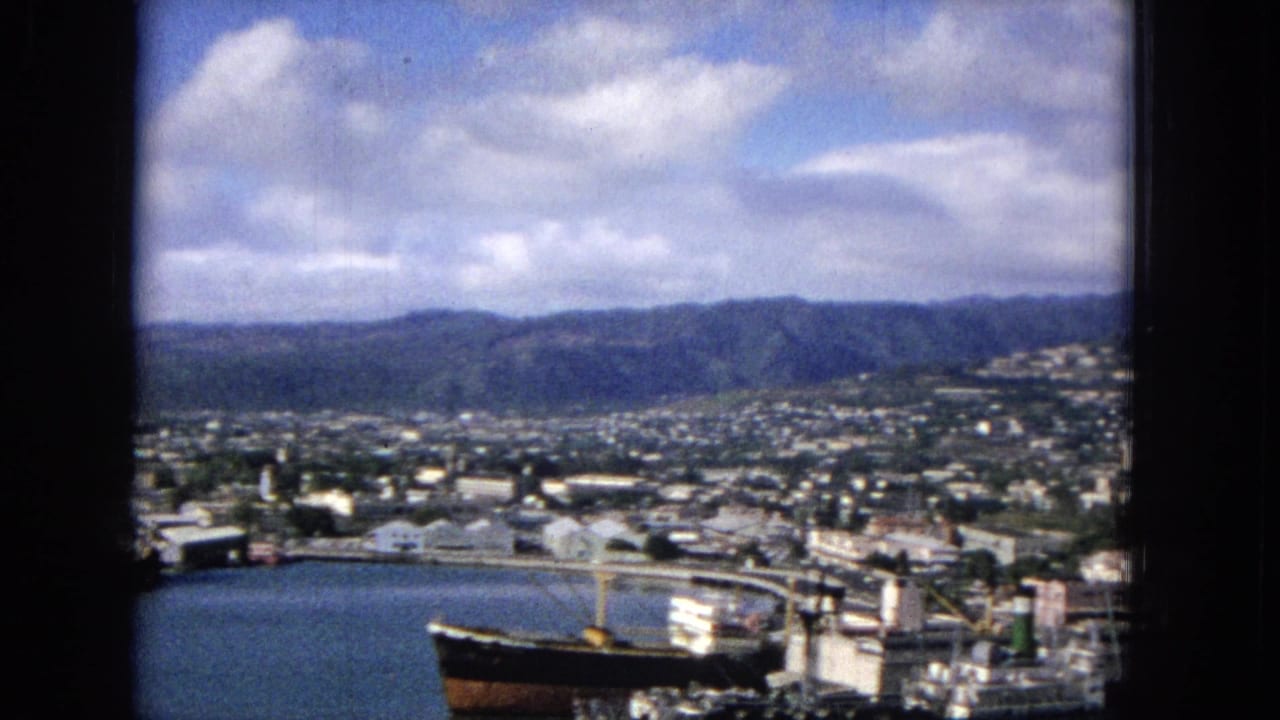 1965 Hawaiian harbor city panorama from an elevated viewpoint, showing bay water, a docked ship, low waterfront buildings, and green mountains under clouds.