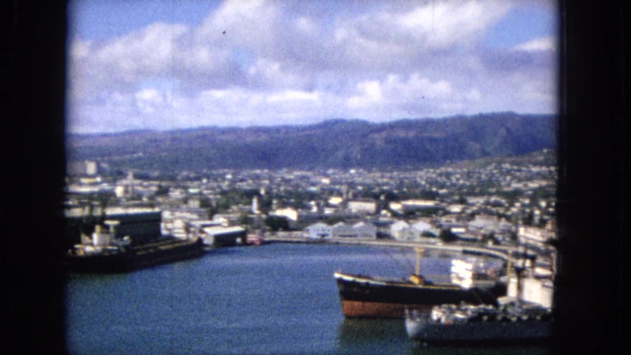 Harbor basin with a large ship, waterfront structures, city buildings, and mountains in the distance.