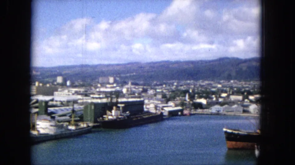 1965 Hawaiian waterfront panorama showing dock areas, low industrial buildings, harbor water, moored vessels, and distant hills in soft home-movie focus.