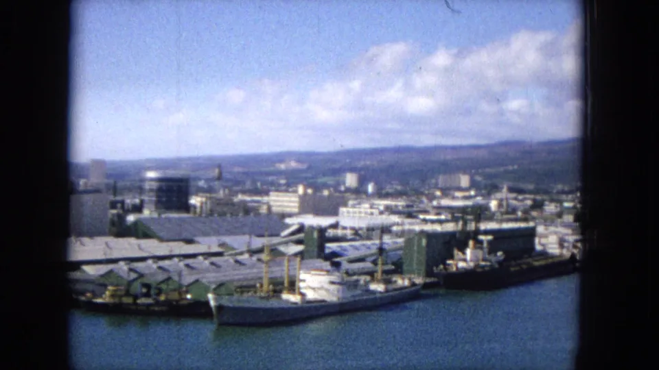 Elevated 1965 Hawaiian harbor panorama with multiple vessels along the waterfront, low pier buildings, city blocks, distant hills, and bright clouded sky.