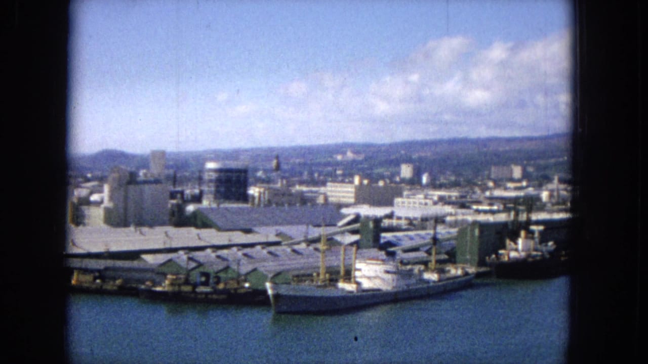 1965 home-movie frame, per metadata, looking across a harbor with docked vessels, long pier roofs, white waterfront buildings, storage tanks, blue water, and distant hills.