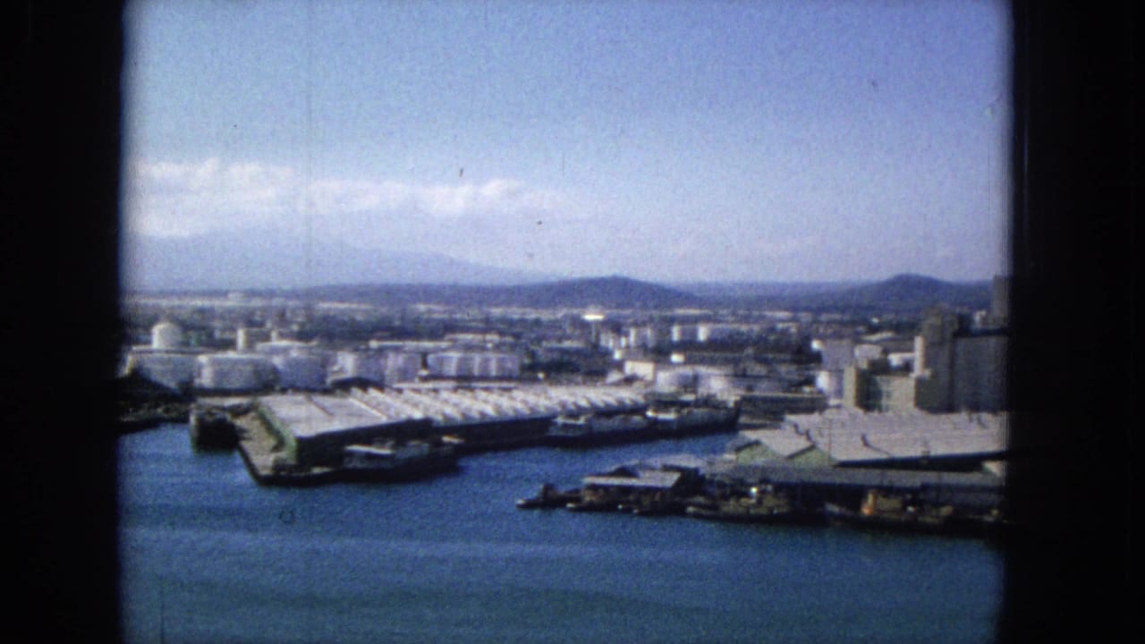 Industrial harbor with piers, storage tanks, docked vessels, water, and low hills beyond the waterfront.