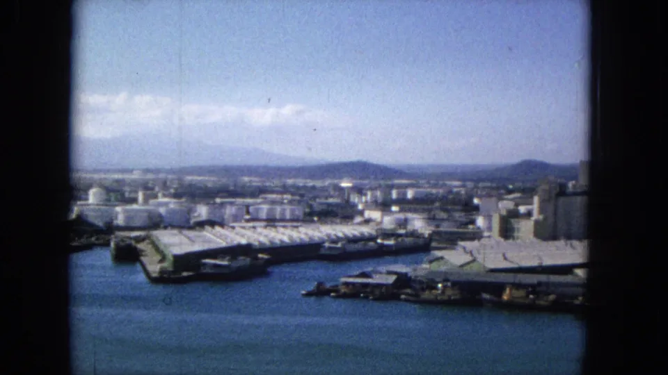 1965 home-movie frame, per metadata, showing a broad harbor view with industrial piers, cylindrical tanks, docked vessels, blue water, distant ridgelines, and a dark vertical frame edge.