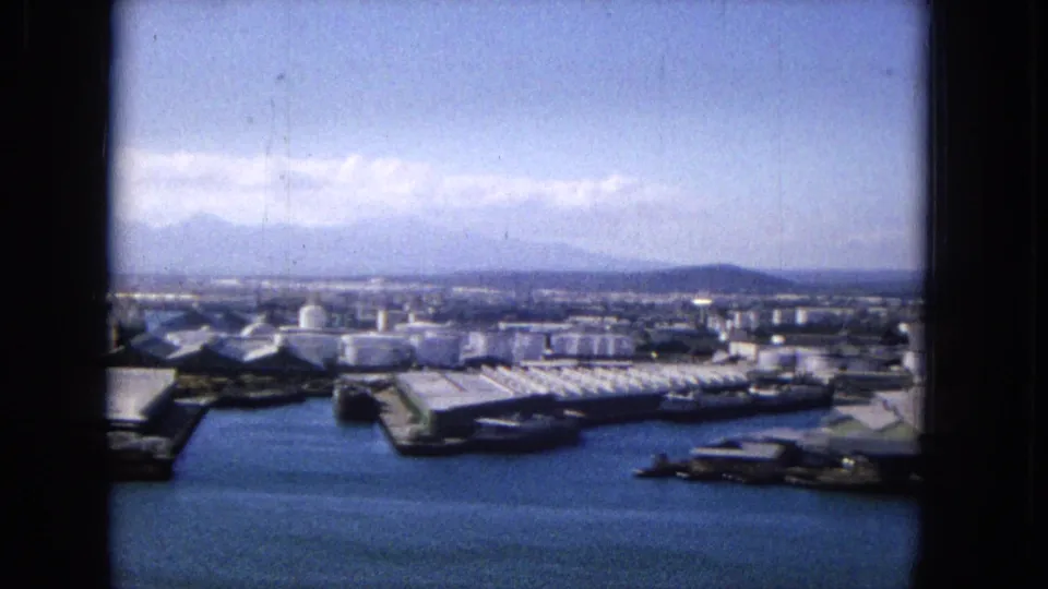 1965 home-movie frame, per metadata, with harbor water below a line of pier roofs, storage tanks, waterfront buildings, and hazy mountain silhouettes under a light cloud band.