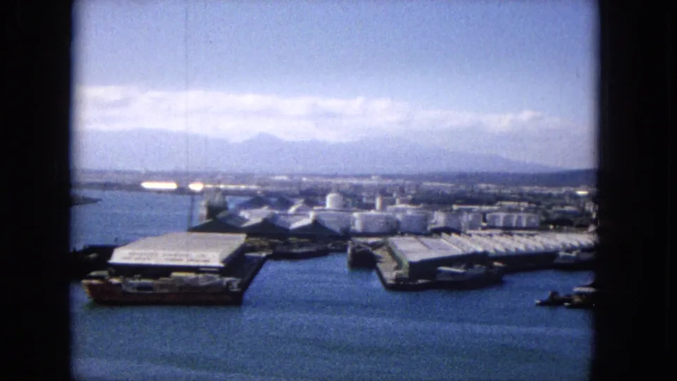 1965 home-movie frame, per metadata, looking over a harbor with blue water, white-roofed waterfront structures, cylindrical tanks, distant mountains, and soft film-frame borders.