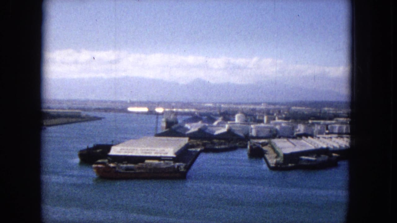 1965 8mm view of a likely Hawaiian harbor with blue water, industrial dock structures, cylindrical storage tanks, low waterfront buildings, and mountains in the distance.