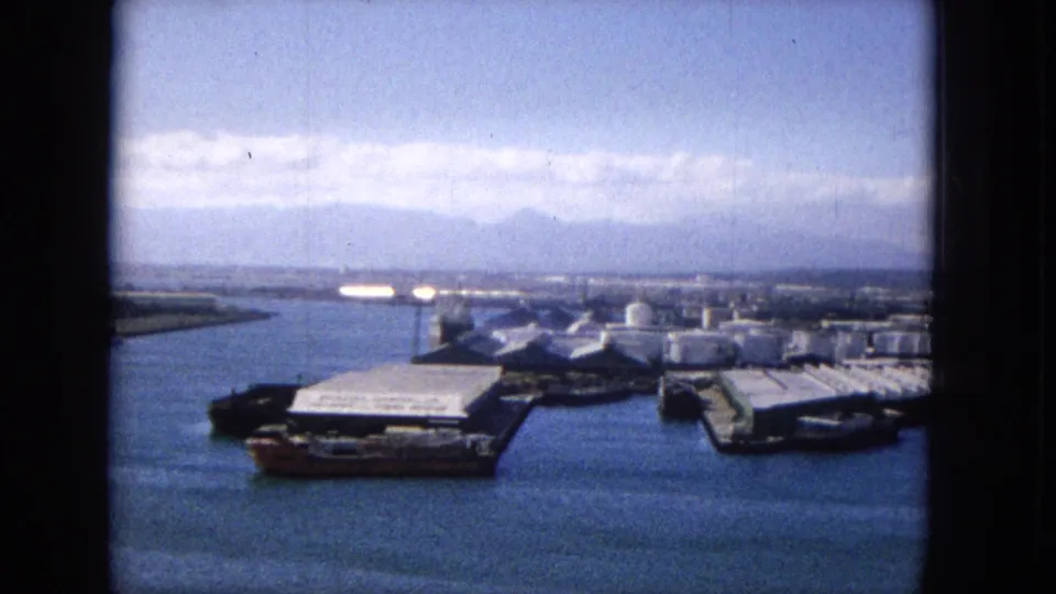 1965 8mm view of a likely Hawaiian harbor with blue water, industrial dock structures, cylindrical storage tanks, low waterfront buildings, and mountains in the distance.