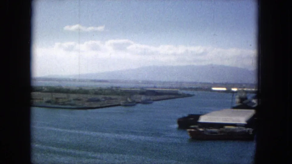 1965 8mm panning view across a likely Hawaiian harbor, with blue water in the foreground, a pier or roofed dock at right, shoreline flats, and distant mountains beneath a bright sky.