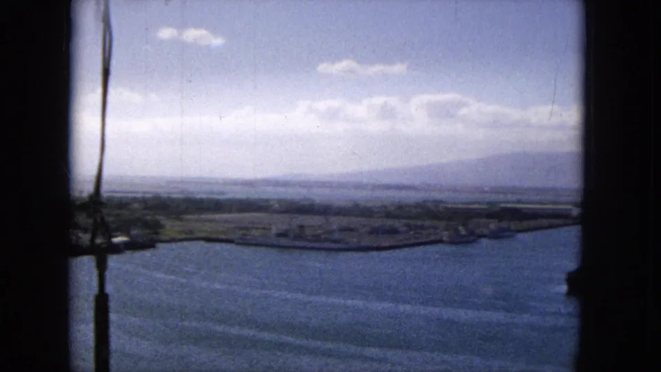 Wide 1965 8mm harbor view, likely in Hawaii, with open blue water, low shoreline, distant mountains, scattered clouds, and a dark film-frame edge at the right.
