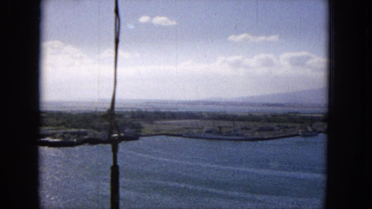 1965 archival footage frame showing an elevated Hawaiian harbor view with water, low shoreline, distant mountains, clouds, and dark vertical framing at the left edge.