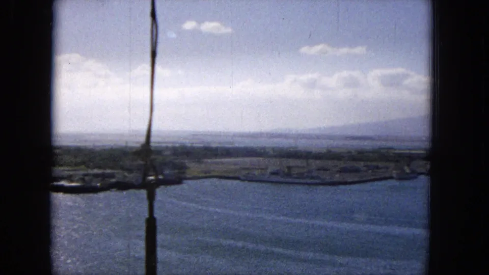 1965 archival footage frame showing an elevated Hawaiian harbor view with water, low shoreline, distant mountains, clouds, and dark vertical framing at the left edge.