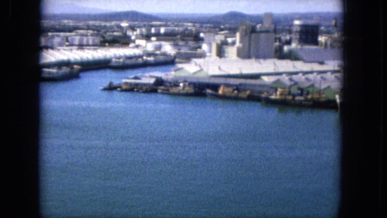 Harbor basin with waterfront buildings, docks, storage structures, and distant hills.