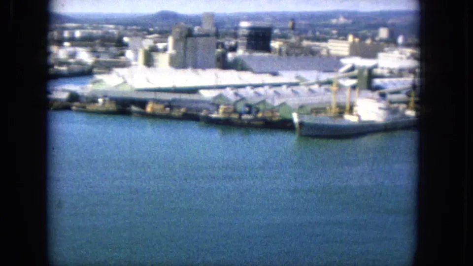 1965 harbor frame showing a large white vessel docked at a pier, with waterfront sheds, tanks, city buildings, and blue water in the foreground.