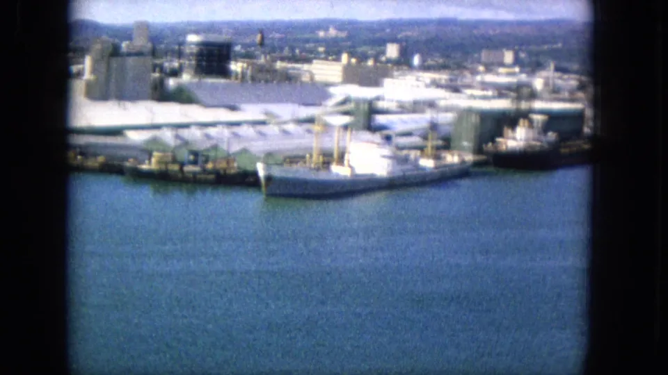 Elevated 1965 Hawaiian harbor view with a white ship at the pier, industrial tanks, low waterfront buildings, distant city structures, and blue water below.