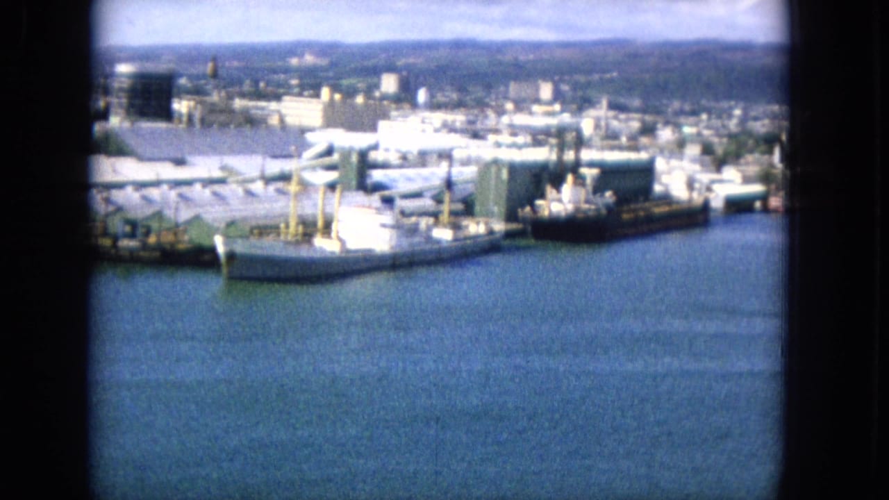 1965 home-movie frame of docked freight ships across blue harbor water, with warehouses, industrial structures, a distant cityscape, and hills beyond.