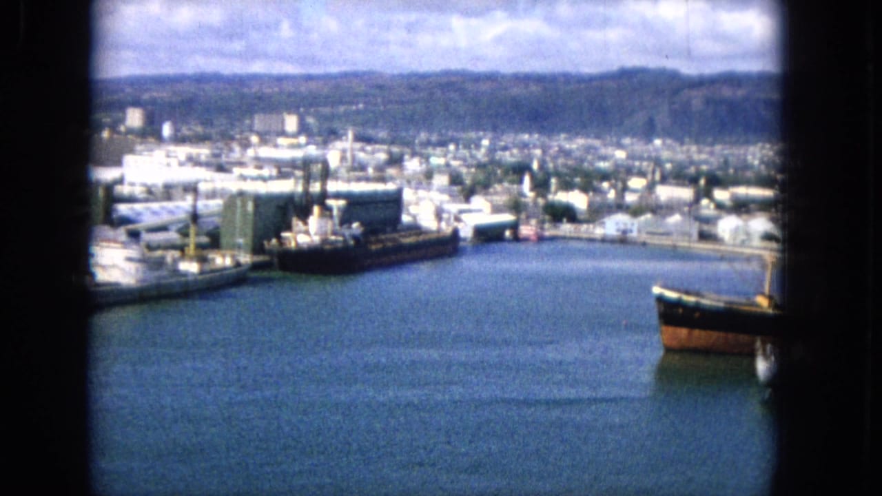1965 8mm frame showing a harbor from above, with docked freight vessels, piers, industrial buildings, city blocks, and hilly terrain under a cloudy sky.