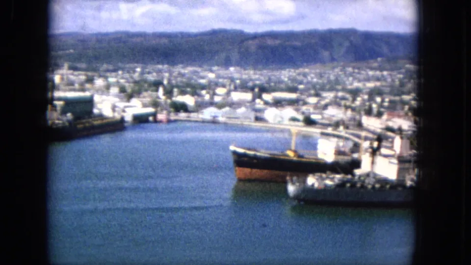 1965 harbor frame with a dark-hulled freight ship near the pier, broad blue water, low waterfront buildings, dense urban development, and hills in the background.