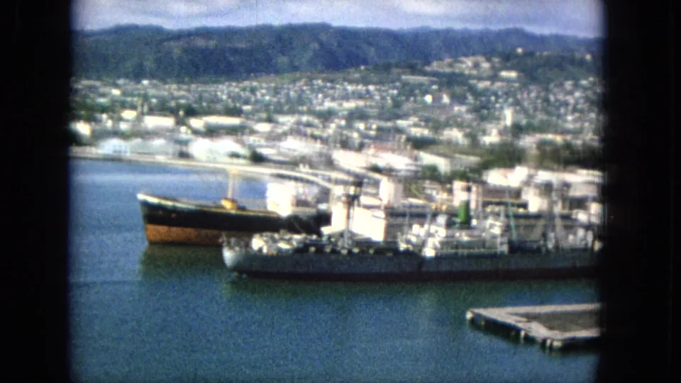 1965 home-movie frame of a harbor with docked vessels, pier equipment, blue water, waterfront industry, a hilly cityscape, and a small floating dock at lower right.
