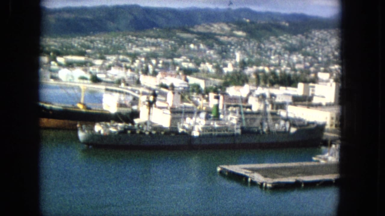 Elevated 1965 home-movie view of a Hawaiian harbor with a large cargo ship at the pier, waterfront buildings, hillside homes, and green mountains beyond.