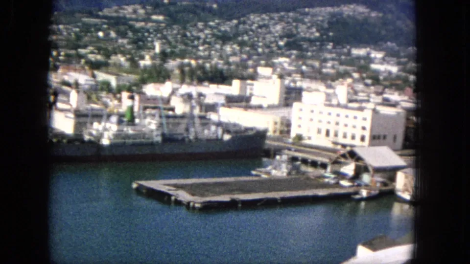 Elevated harbor view from the 1965 home movie, showing a cargo ship near the waterfront, a pier, pale buildings, hillside housing, and mountain slopes.