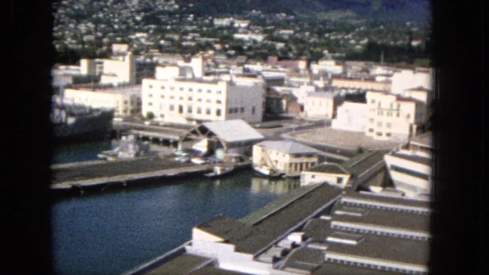 The pan moves across the Hawaiian port city waterfront, revealing piers, low-rise buildings, harbor water, and residential neighborhoods climbing the hillside.