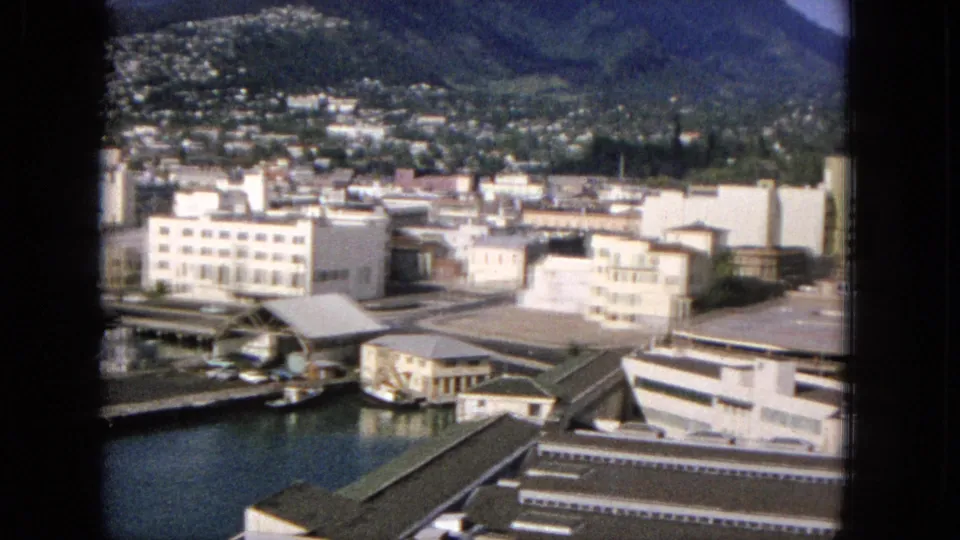 Final frame of the 1965 harbor pan showing low waterfront buildings, dock structures, water in the foreground, dense hillside residences, and mountains behind the city.