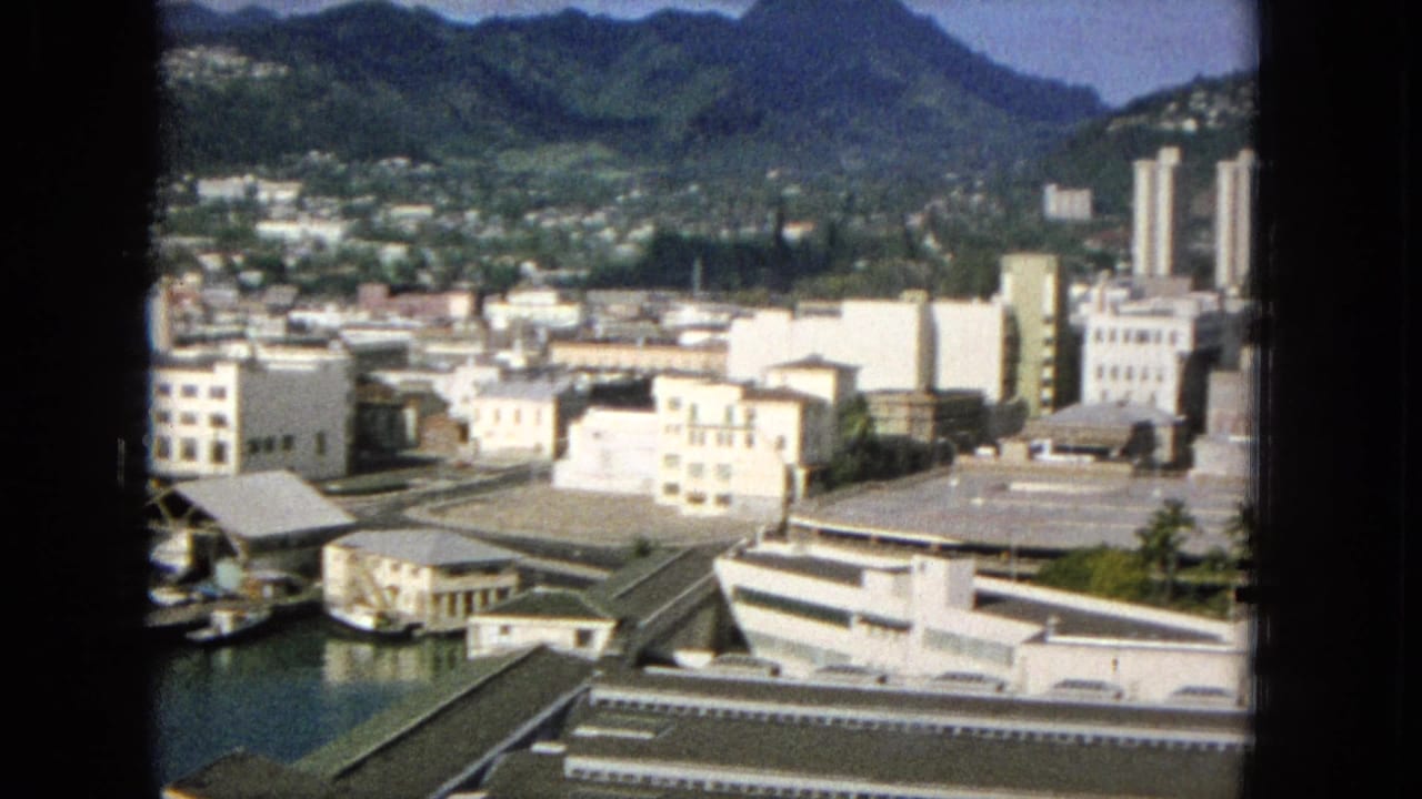 1965 Super 8 frame showing a Hawaiian city panorama from an elevated window, with waterfront structures, low-rise buildings, roads, greenery, and mountains in the distance.