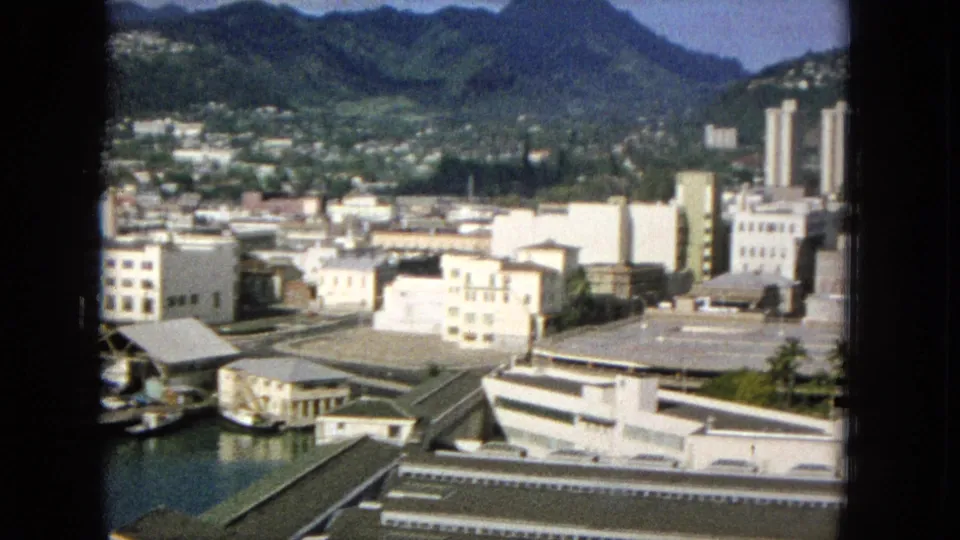 1965 Super 8 frame showing a Hawaiian city panorama from an elevated window, with waterfront structures, low-rise buildings, roads, greenery, and mountains in the distance.