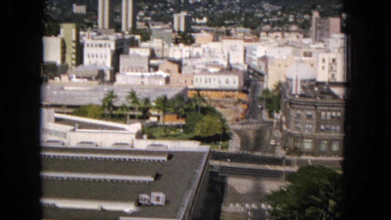 Elevated 1965 Super 8 view across a Hawaiian urban district, showing pale mid-century buildings, roads, trees, a large older block, and hillside development in the background.