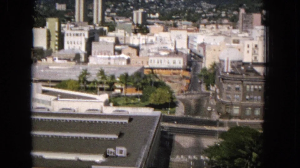 Elevated 1965 Super 8 view across a Hawaiian urban district, showing pale mid-century buildings, roads, trees, a large older block, and hillside development in the background.