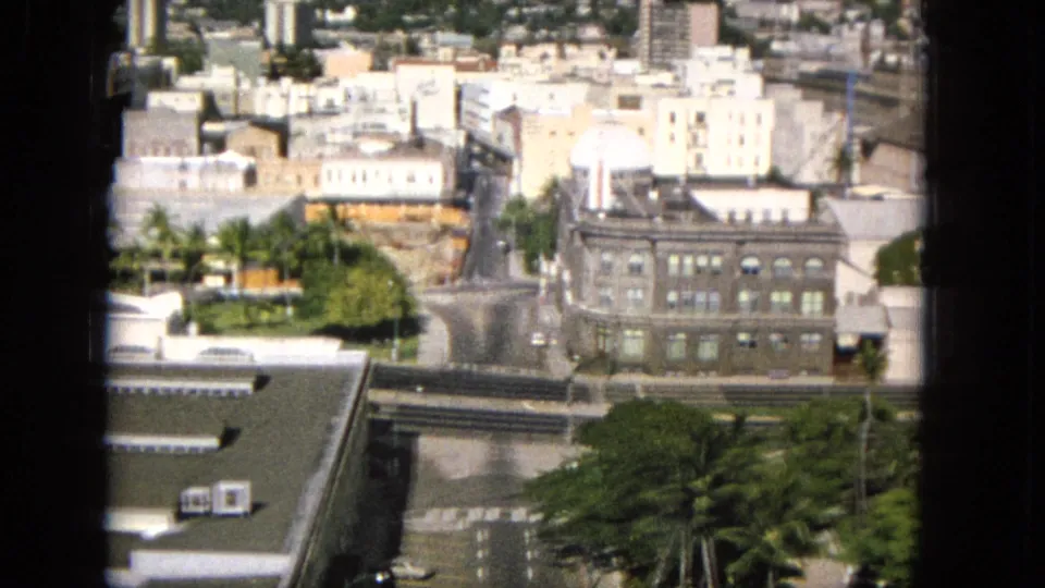 1965 Super 8 frame looking down over a Hawaiian city district with a large masonry building, pale rooftops, roads, trees, parked cars, and the edge of a modern roof in the foreground.