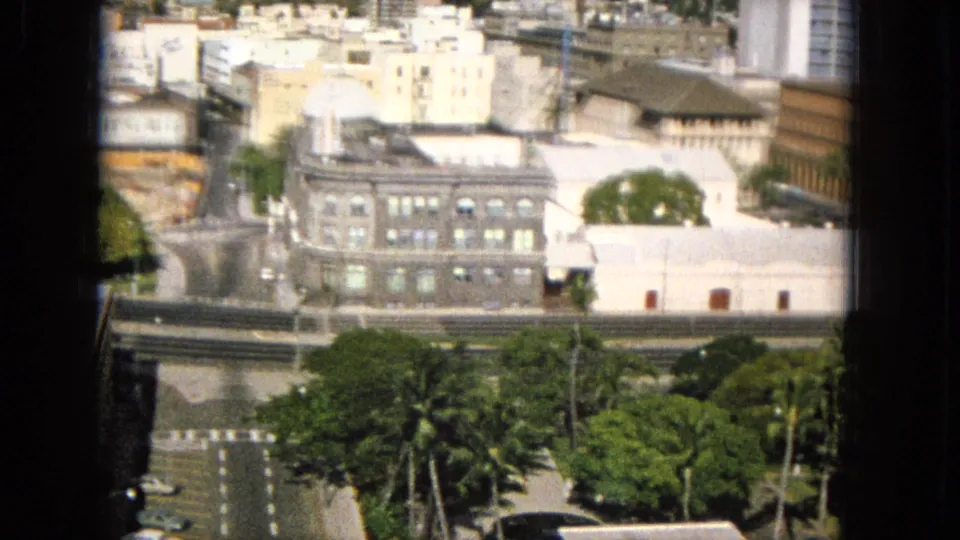 1965 Super 8 frame of a Hawaiian city view through a dark window border, with a large masonry building, intersecting roads, trees, parked cars, and bright low-rise structures nearby.