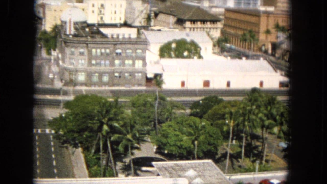 1965 8mm frame likely showing Honolulu from an elevated interior or balcony vantage, with palms, a multi-lane road, low civic-looking buildings, and dark window mullions in front.